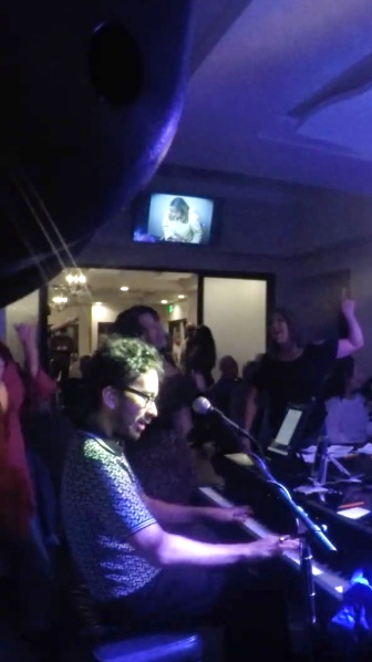 Steven at the piano during a dueling pianos show, second pianist and crowd behind him